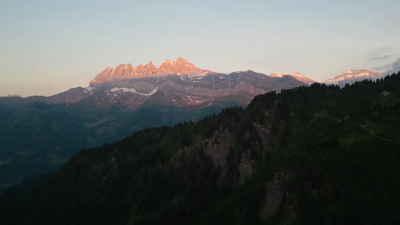 Beautiful Sunset drone flight in the Swiss Alps. Drone pans behind the tree line looking at the pink sunset on the peaks of the Dents du Midi in the Val d'Illiez of Canton Valais in Switzerland.