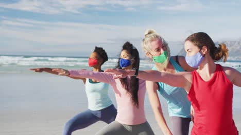 group of diverse female friends wearing face masks practicing yoga at the beach