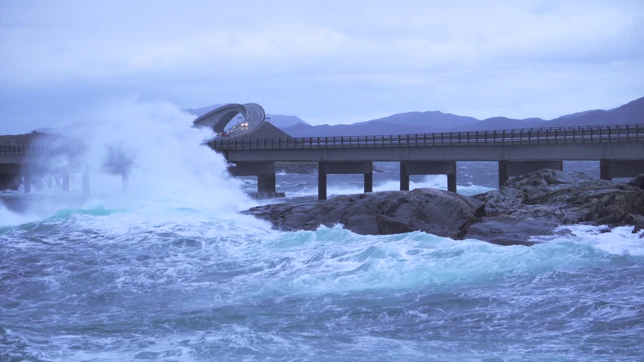 Beautiful scenery of waves crashing into rocks next to a bridge with cars driving on it.
