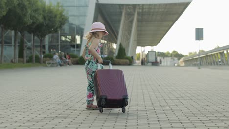 Child-girl-tourist-with-suitcase-near-airport.-Kid-wave-hand,-walks-down-street-with-bag.-Vacation