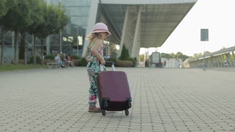 child girl tourist with suitcase near airport. kid wave hand, walks down street with bag. vacation