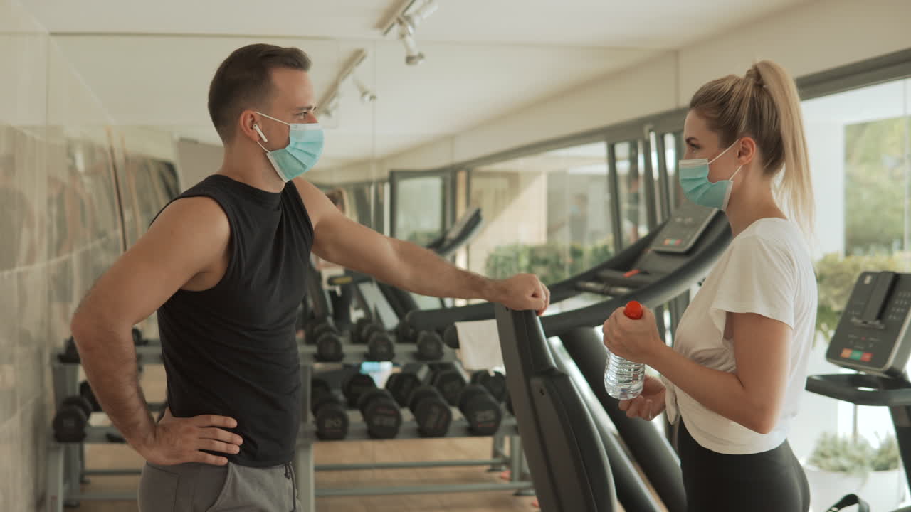 젊은 운동선수 여성과 얼굴 마스크를 쓴 남자가 체육관에서 이야기를 나누고 있습니다. 코로나19 대유행 기간 동안 스포츠.
