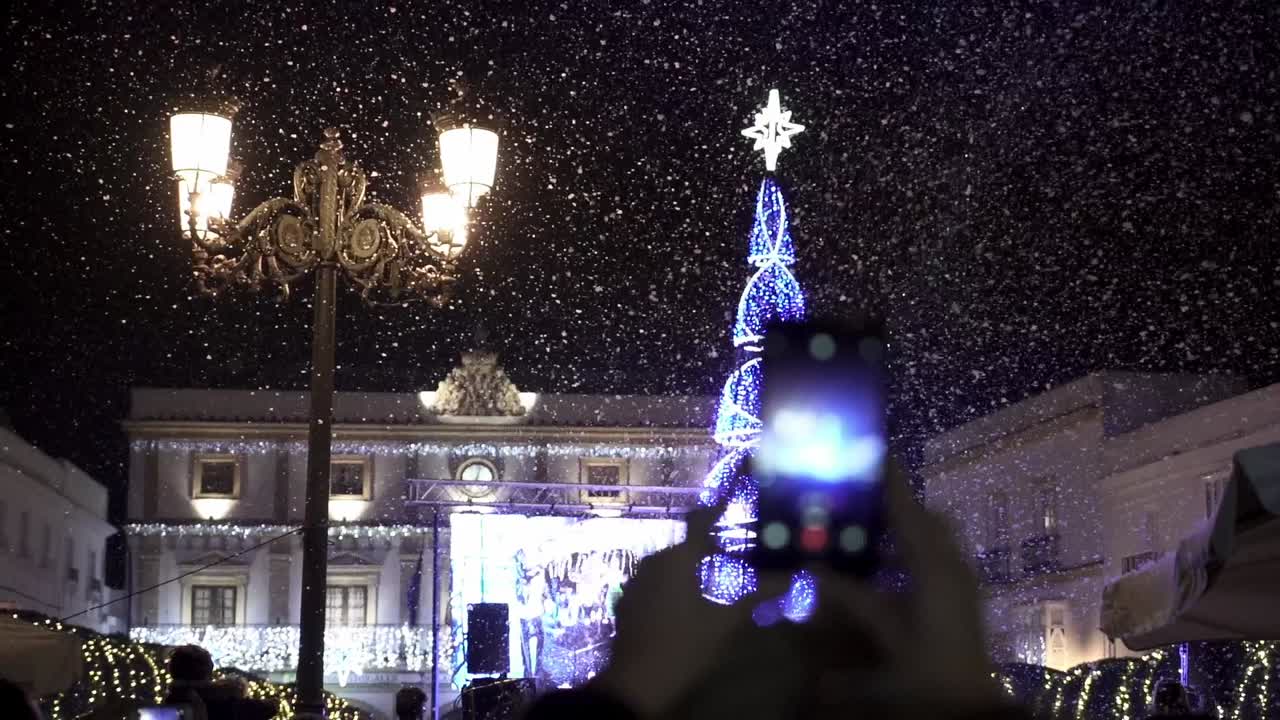 스마트폰을 가진 사람이 강설량으로 촬영하는 동안 별이 위에 있는 조명이 켜진 크리스마스 트리가 있는 카디스 스페인 메디나 시도니아의 스페인 크리스마스 파티의 슬로우 모션 핸드헬드 샷