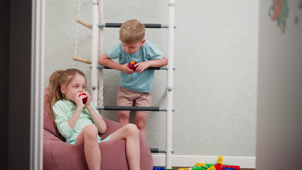 children fresh fruit i eating indoors, girl relaxing on pink bean bag eating apple while boy hangs on indoor ladder also eating fruit, colorful toys scattered on wooden floor adding playful vibe