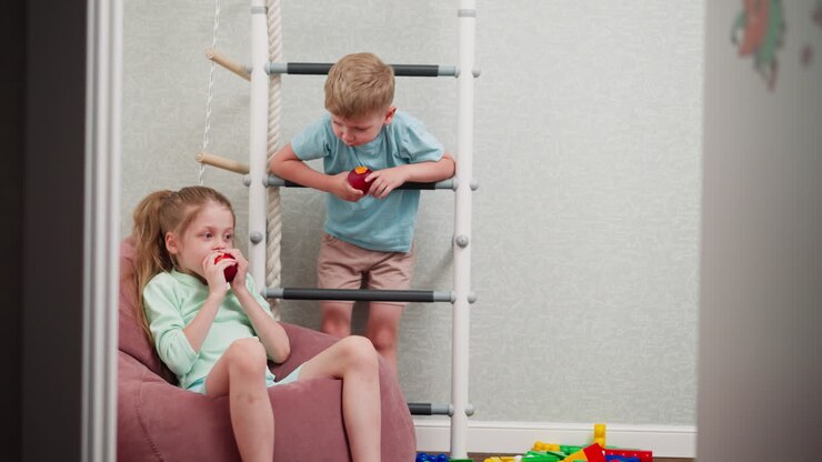 children fresh fruit i eating indoors, girl relaxing on pink bean bag eating apple while boy hangs on indoor ladder also eating fruit, colorful toys scattered on wooden floor adding playful vibe