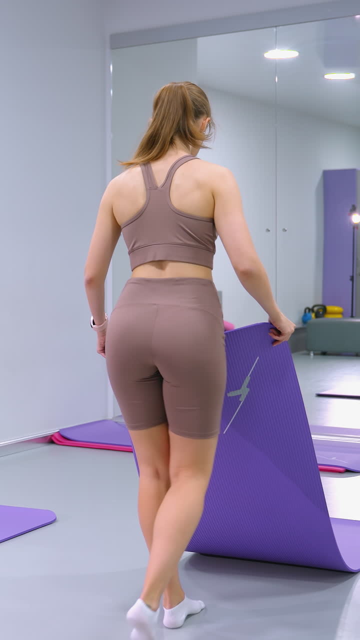 mujer deportiva recoge una alfombra de yoga para arreglar con otros en un elegante estudio de fitness, un gran espejo refleja el entorno, mostrando un espacio limpio y organizado con equipos de ejercicio