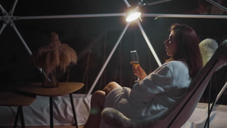 mujer sombría sosteniendo un vaso en las manos distraída juguetea con el objeto que acompaña a sus propios pensamientos. calor no forzado del silencio noche envolve en el sillón