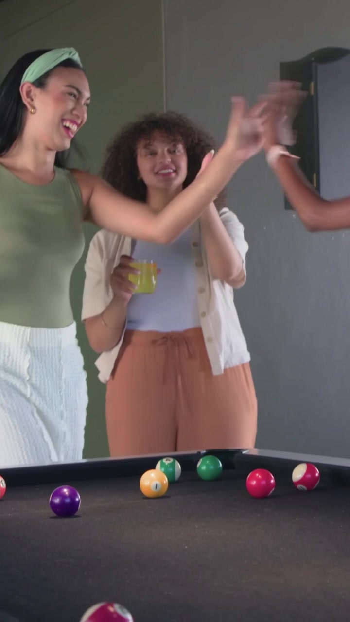 Video verticale: Panoramica su una donna riccia con un drink, saluta una voce fuori campo al tavolo da biliardo, indossa un top bianco.