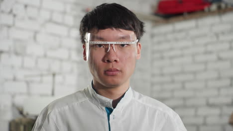 young lab technician wearing lab coat and protective glasses nods head continuously, reflection seen on protective glasses adds depth, background features brick wall and red object