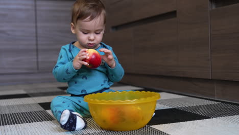 lovely toddler sits on the floor with a yellow bowl in front of him. cute kid takes an apple from a dish, looks at it carefully and turns it in his hands.