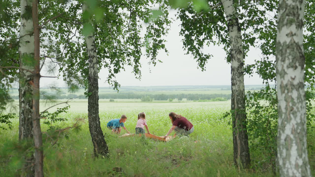 자작나무 아래에서 흰색 옷을 입은 가족이 피크닉을 즐기고 있어요. 엄마는 무릎을 꿇고 담요를 깔고, 두 아이는 키 큰 풀밭에서 놀고 있어요. 멀리에는 호수와 탁 트인 하늘이 보이고, 부드러운 바람과 따뜻한 여름 햇살이 느껴지는 자연스러운 시골 풍경이에요.