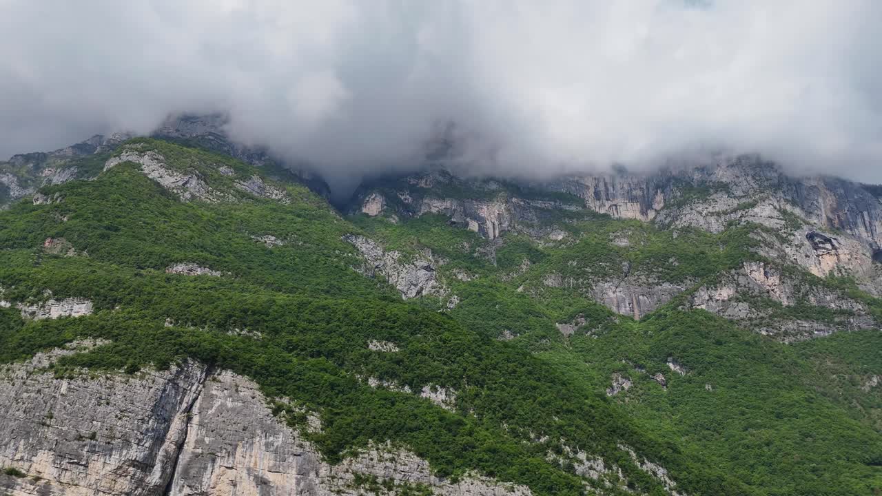 Aerial footage of the Italian Dolomites, showing steep rocky terrain, dense greenery, and a misty valley under cloudy skies