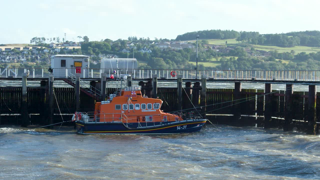 Orange lifeboat approaches wooden pier on choppy water, daylight, wide shot, static camera