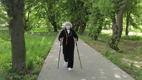 Active-senior-old-woman-in-mask-training-Nordic-walking-in-park-at-quarantine