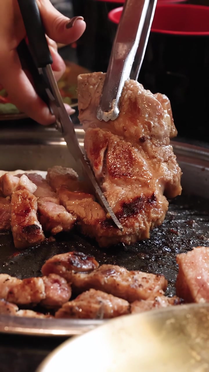 Tangan memotong daging babi panggang dengan penjepit dan gunting di atas panggangan barbekyu Korea meja, pencahayaan hangat