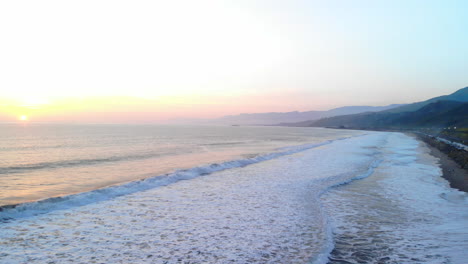 Aerial-shot-of-ocean-waves-crashing-on-the-beach-under-a-colorful-sunset-sky-background-along-the-coast-of-Ventura,-California-DRONE