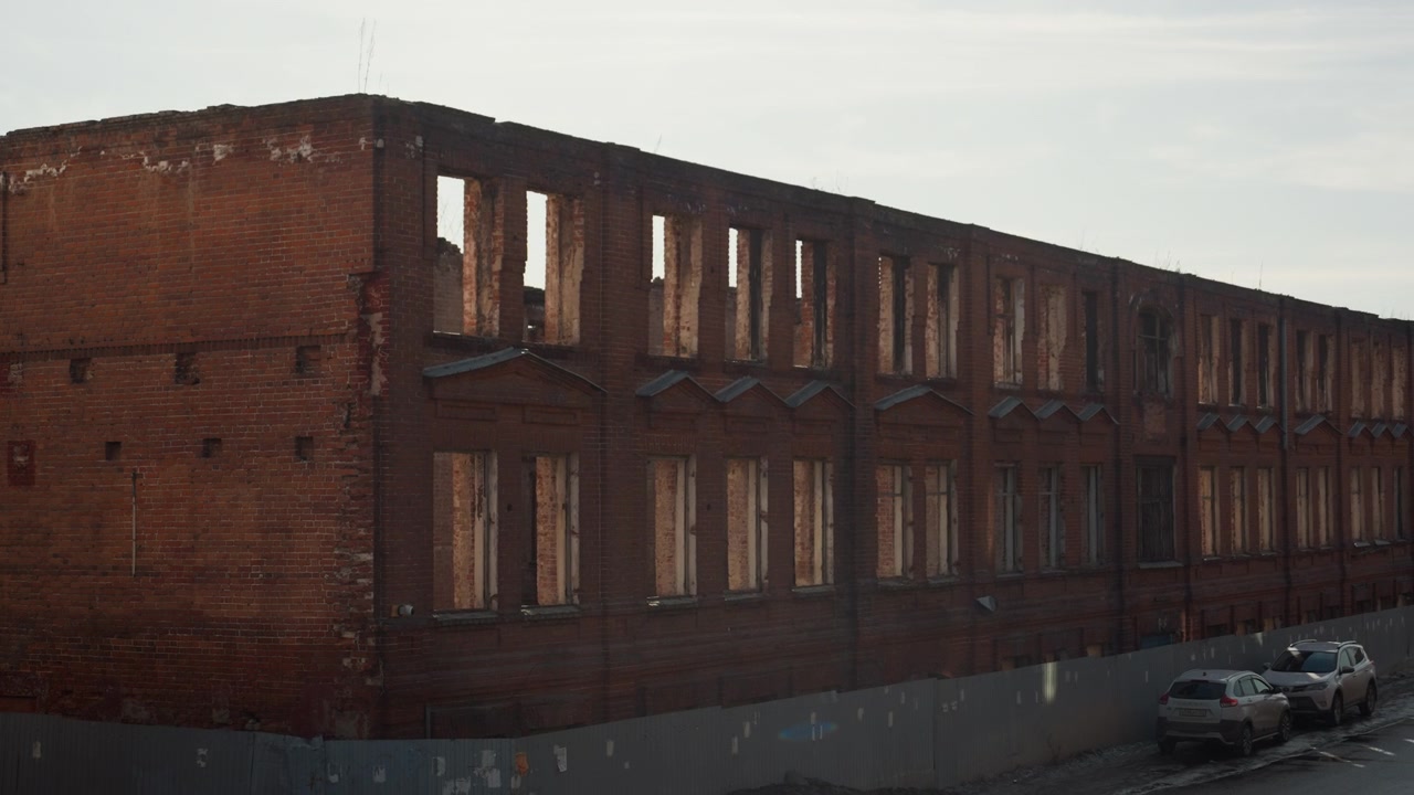 Bel magazzino in mattoni al tramonto, finestre vuote che brillano di un caldo bagliore, lunga facciata in muratura rossa, auto parcheggiate su pavimentazione screpolata, cielo coperto e cupo, luce cinematografica soffusa, tranquilla desolazione mista a elementi storici.