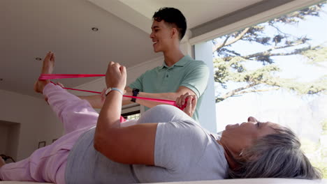 helping senior woman with leg exercises, male physiotherapist using resistance band
