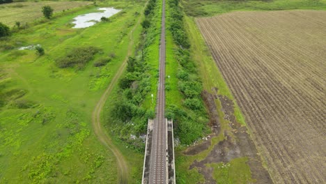 aerial footage over an elevated railway revealing a farm on the right, grassland on the left and a ponds, in saraburi, thailand