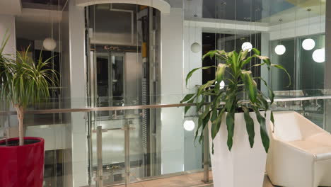 a modern indoor setting featuring potted plants positioned on either side of a glass elevator. there are two red pots, two white pots, and one brown pot with tall green plants