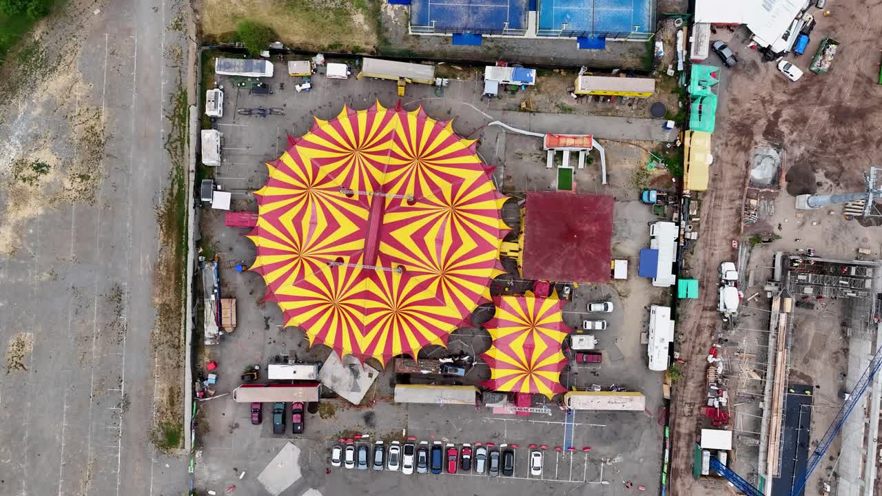 Ascending top-down drone aerial of colorful striped circus tents with carnival setup, vehicles, trailers, and backstage area overview