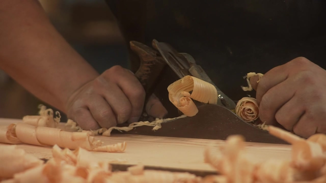 Close up of craftsman planing wooden board with hand plane, producing curled wood shavings while smoothing timber surface on workbench during woodworking