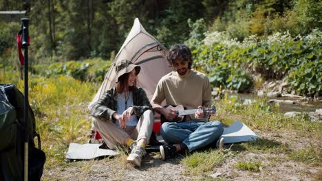 a man and a girl tourists are sitting near their tent, the man plays the guitar and the girl dances and sings