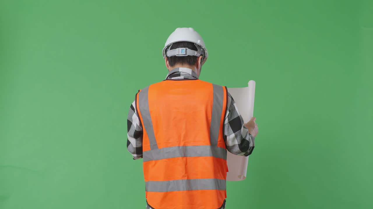 vista trasera de ingeniero masculino asiático con casco de seguridad mirando el plano en sus manos y mirando a su alrededor mientras camina en el estudio de fondo de pantalla verde