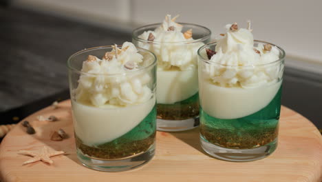 round view of dessert-like candle in transparent glass with layered green base, creamy middle, and whipped topping garnished with tiny shells, placed on wooden table with scattered sea-themed decor