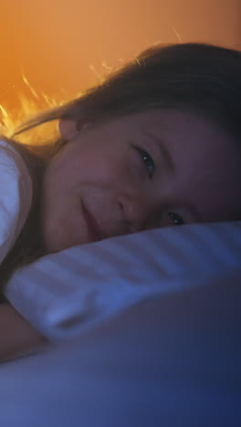 little girl smiles looking into camera under soft blanket on comfortable bed in dark room partially illuminated by night lamp in toy teepee at home slow motion