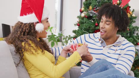 Video-of-happy-diverse-couple-exchanging-christmas-gift-and-embracing-at-home
