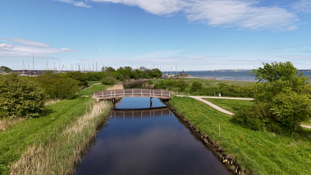 Aerial drone view of a calm reflective canal bordered by green fields, reeds, and scattered trees under a bright blue sky