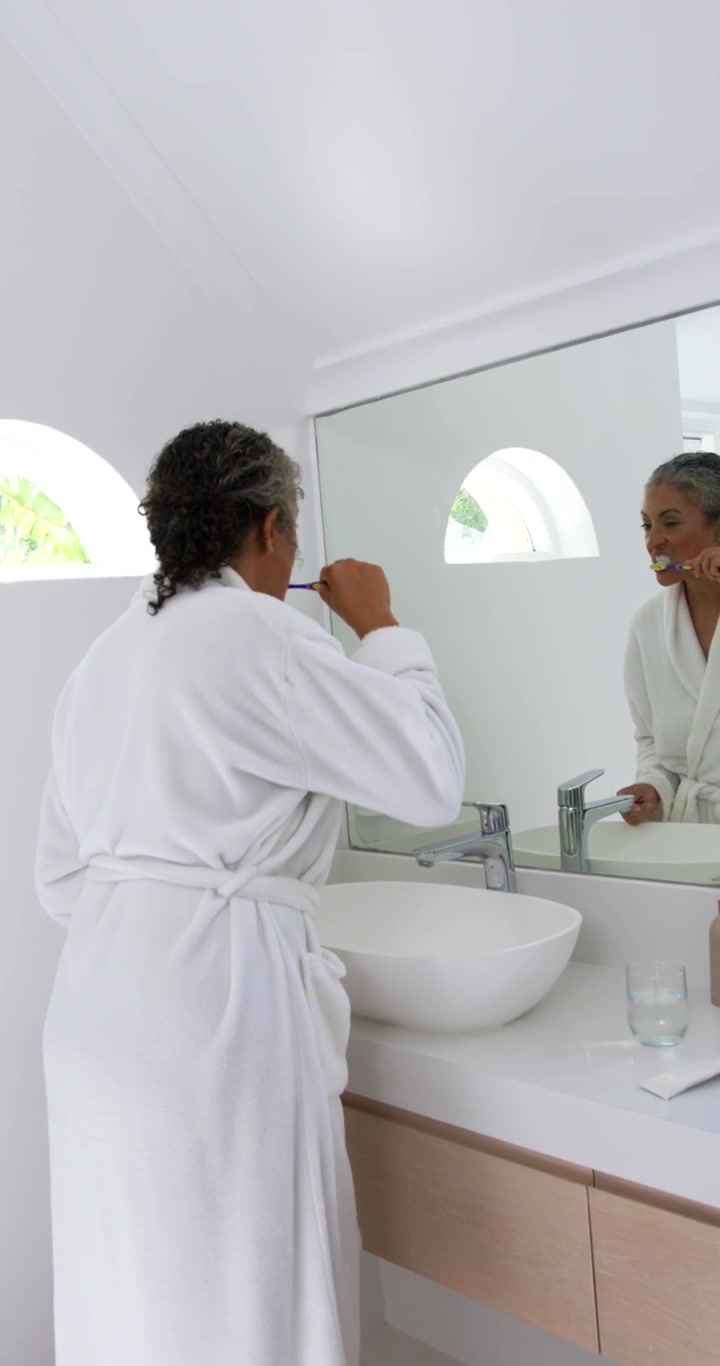 African woman in robe holding toothbrush near mouth, brushing teeth at vessel sink for morning care