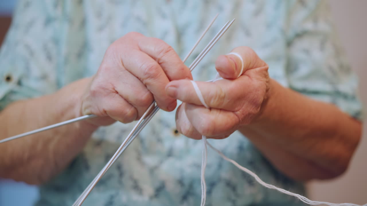 Closeup elderly white grandma knitting, steady fingers working thin metal needles and soft yarn, floral blouse visible, warm indoor light, intimate nostalgic mood, skilled textile craft and patient