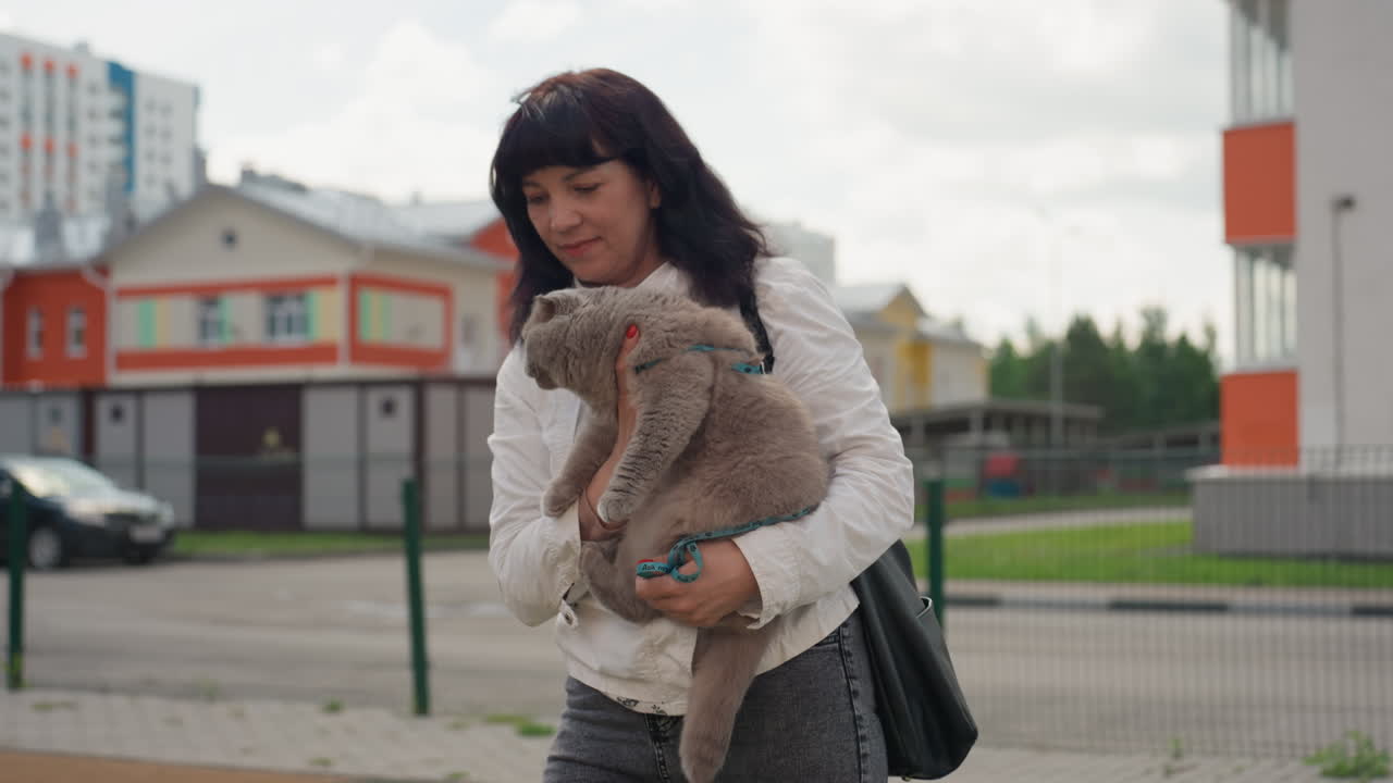 Female Individual Takes Pet For Walk Near Homes, Woman And Her Cat Enjoy Casual Stroll In Park Area, Woman Bends Down To Allow Her Cat To Explore While Walking Near Urban Residences