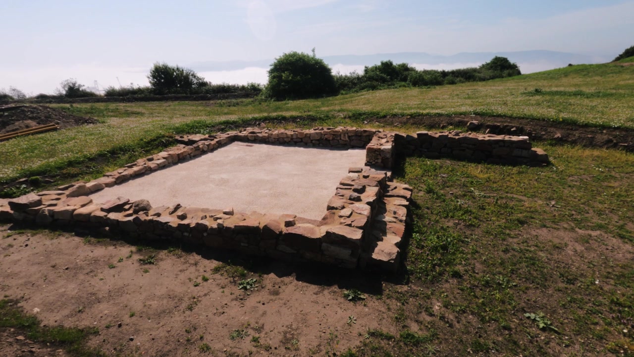 Exposed stone foundation ruins at La Campa Torres archaeological park in Gijón, Asturias. Green hillside and coastal haze in background.