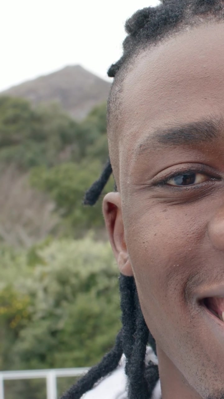 Video verticale: Uomo sorridente con dreadlocks, con maglietta chiara, che sbatte le palpebre dopo aver guardato, muovendosi a destra sul ponte, vicino alla ringhiera.