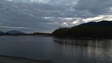 river, shore with trees and mountains at sunset. spring season. fraser valley, british columbia, canada.