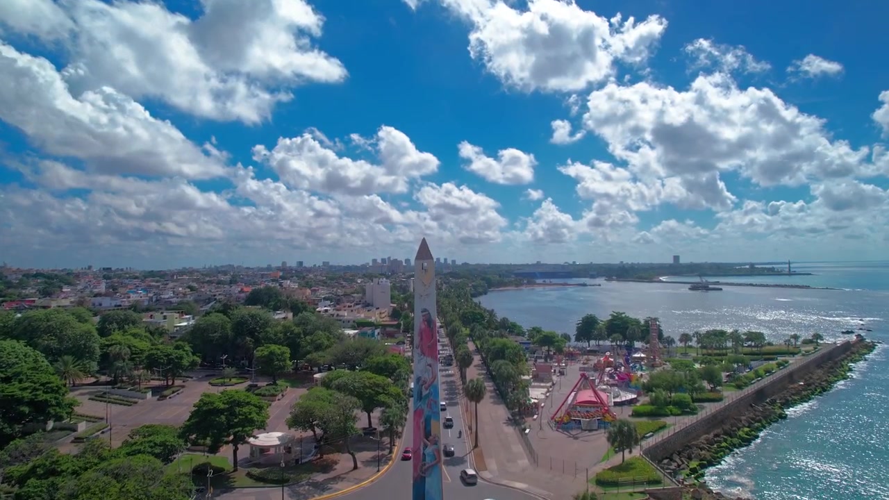 Tomada aérea de drone avançando e subindo sobre o monumento Obelisco Hembra na Avenida George Washington para revelar a costa caribenha, o mar e o horizonte da cidade em Santo Domingo