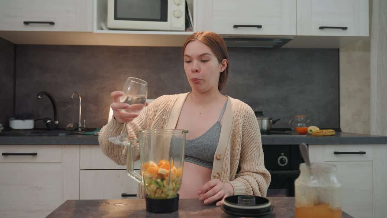 Donna incinta in cucina che versa acqua su mandarini sbucciati e kiwi a fette, che indossa un cardigan comodo e un bralette, mattina tranquilla, frullatore da banco, che prepara un delicato frullato prenatale, mani in primo piano