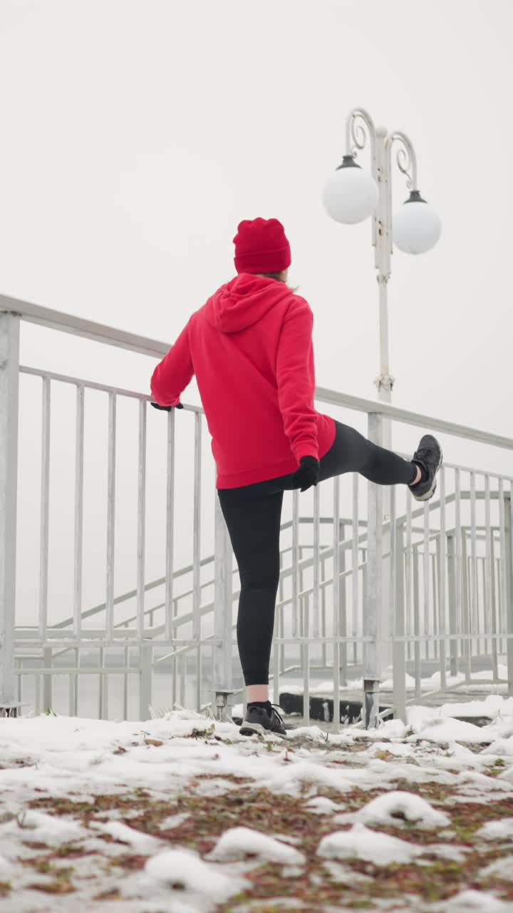 athlete exercising outdoors during winter holding iron railing for support while stretching leg back and forth amid foggy atmosphere with light poles stairs snowy ground and river background