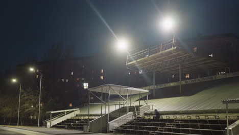 a man dressed in black and wearing a jacket sits quietly in an empty stadium at night, with a soccer ball by his side, the background is illuminated