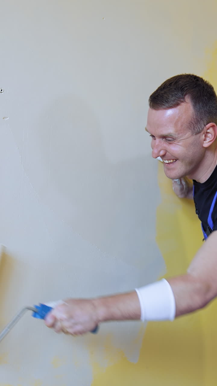 Positive worker doing renovation in the room. Smiling builder painting wall in the light color with a paint roller. Room makeover. Vertical video