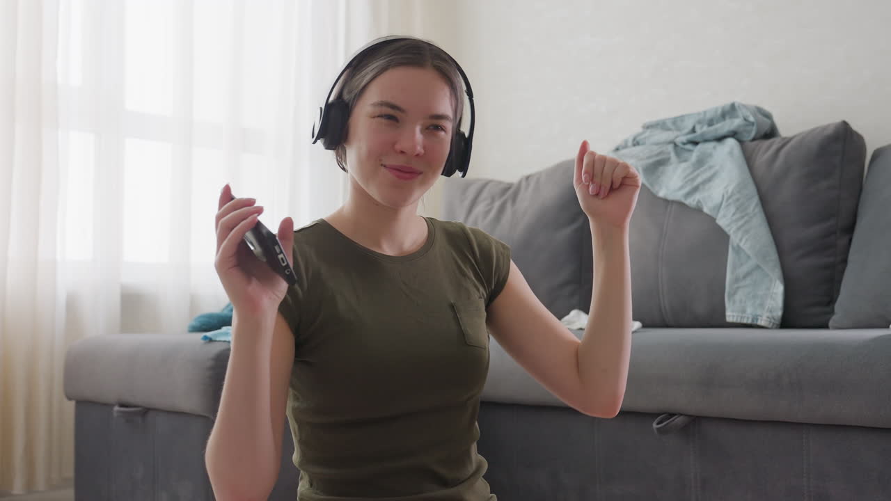 Young woman seated indoors wearing headphones dancing with joyful expression while holding phone in hand, casual clothes on sofa background