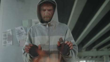 homeless man warming hands by a fire barrel under a bridge