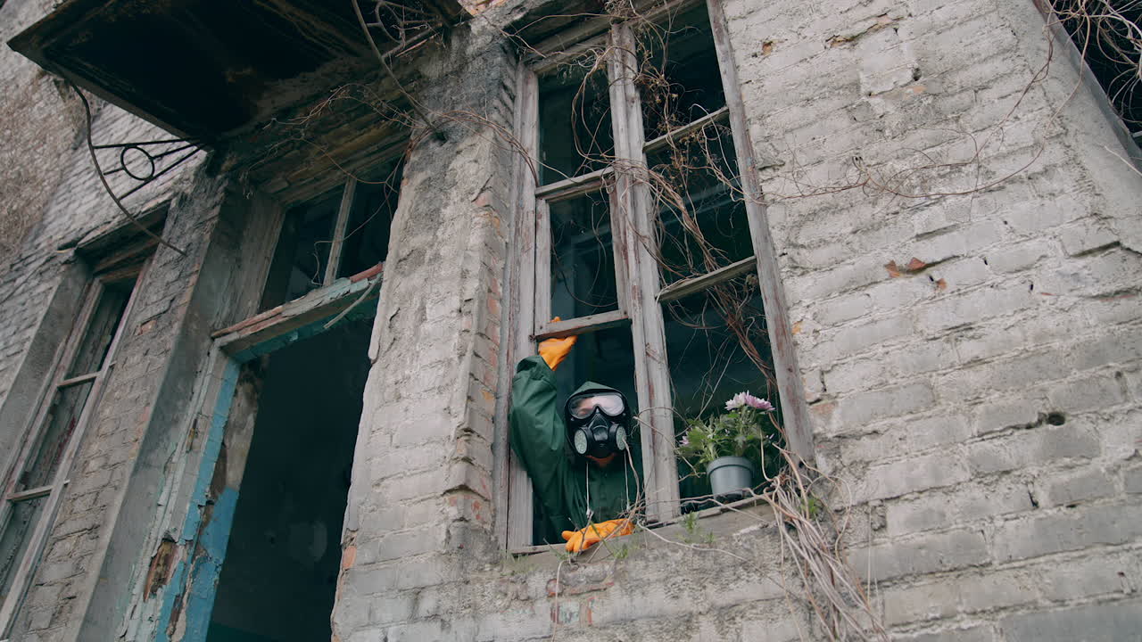 Ecologist wearing respirator alone in empty house. Man in safety suit and mask looking out of the window without glass from abandoned building. Chemical attack.