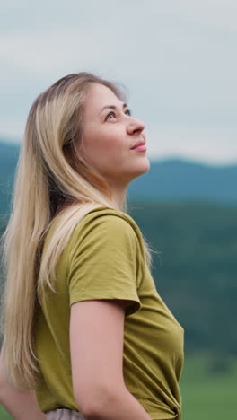 positive woman hiker looks around holding hands in pockets against mountains with green slopes at highland reserve on overcast day slow motion