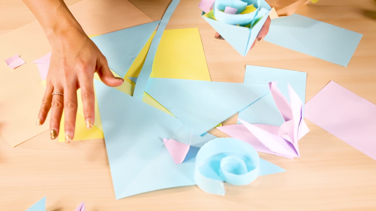 Person arranges colorful origami flowers and paper shapes under bright, even lighting, overhead view