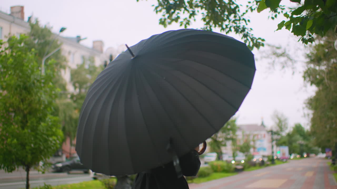 Woman in long black coat adjusting black umbrella outdoors on city street with green trees and buildings, overcast sky, autumn atmosphere, urban lifestyle moment with thoughtful expression