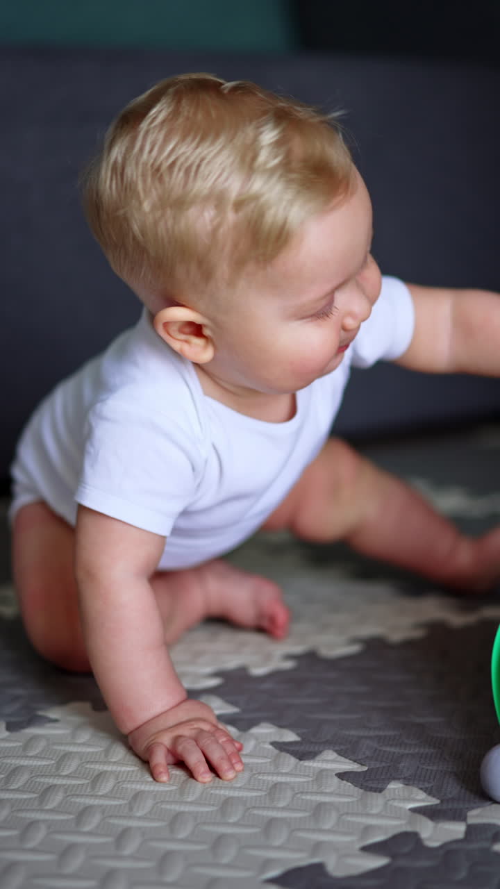 Adorable blond Caucasian blue-eyed baby sitting on the floor. Close up portrait of a cute child wearing white bodysuit. Vertical video.